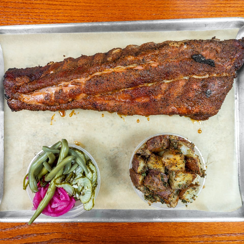 Smoked barbecue ribs served on a tray with seasoned potatoes and pickled green beans.