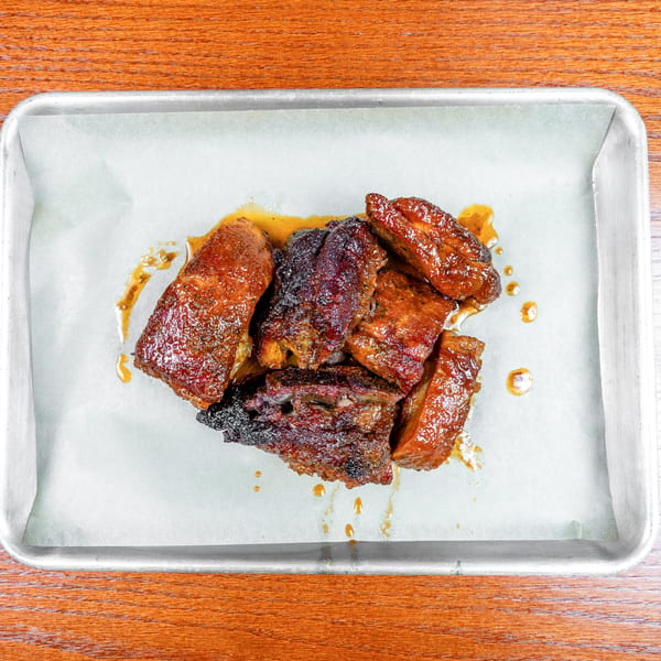 Glazed, smoked meat pieces piled on parchment in a metal baking tray atop a wooden table.