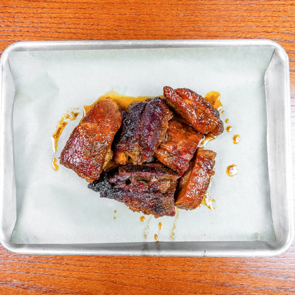 Glazed, smoked meat pieces piled on parchment in a metal baking tray atop a wooden table.