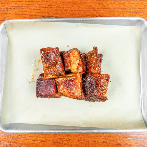 Glazed, caramelized meat cubes arranged on parchment in a metal baking tray atop a wooden table.