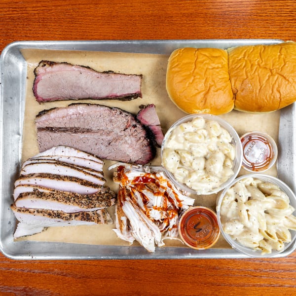 Overhead tray of sliced barbecue brisket, turkey, pulled pork, buns, macaroni salad, and barbecue sauce on a wooden table.